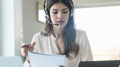 Une femme avec un casque d’écoute de téléphone qui regarde des papiers et son ordinateur portable.