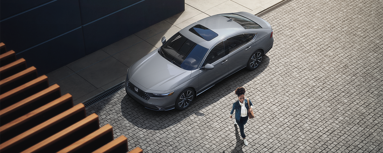 3/4 bird’s eye view of woman walking away from a grey Accord parked curbside in an upscale urban setting.