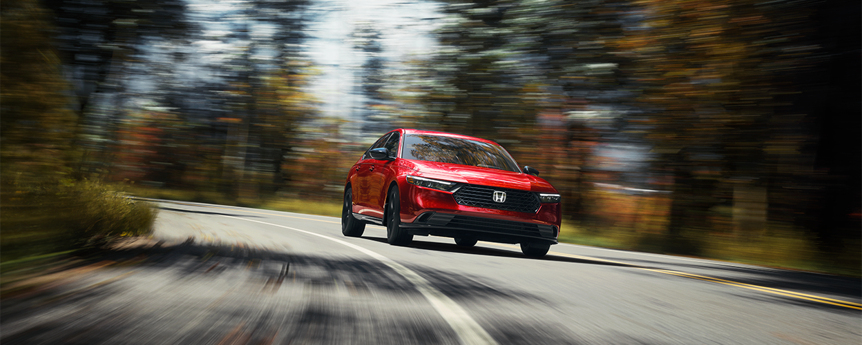 3/4 front view of red Accord, driving toward camera, taking a turn on a temperate rainforest highway in the day.