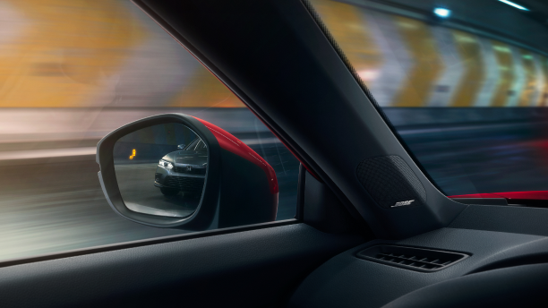 Interior closeup of the door mirror with the reflection of a car in it and a lit up blind spot indicator light.
