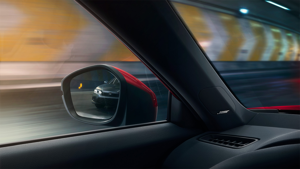 Interior closeup of the door mirror with the reflection of a car in it and a lit up blind spot indicator light. 