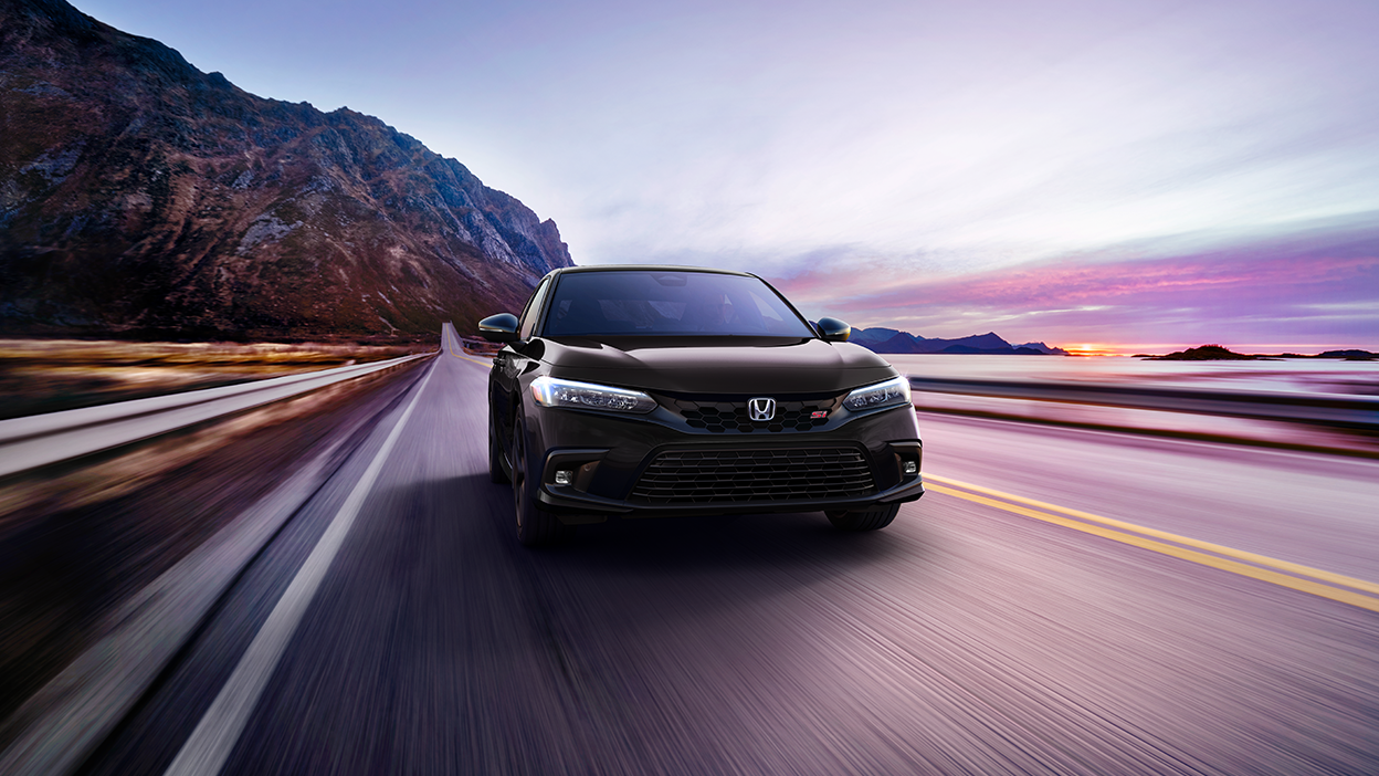 Front view of black Civic Si driving toward camera on a mountainous oceanside highway.