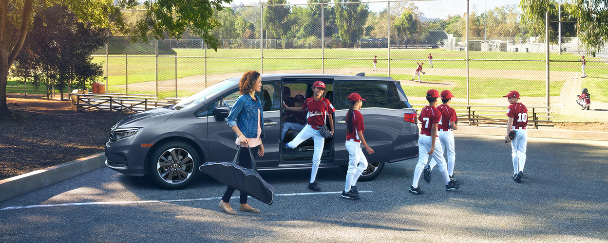 Mom and kids’ baseball team exiting a grey Odyssey parked near a baseball diamond.