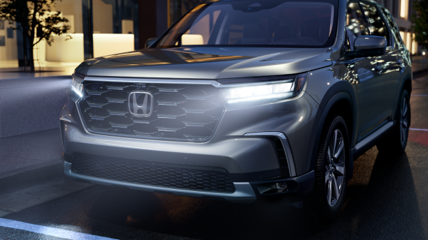 3/4 front view of grey Pilot, parked curbside on a city street with front headlights on.