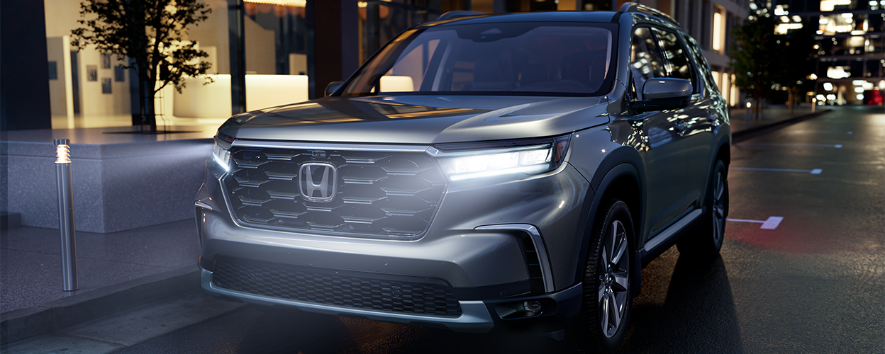 3/4 front view of grey Pilot, parked curbside on a city street with front headlights on.