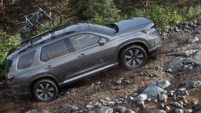 Wide sideview of grey Pilot, with a bike on the roof rack,driving uphill on a dirt road in a temperate rainforest. 